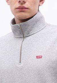 Εγγύς λήψη ατόμου που φοράει ανοιχτό γκρι πουλόβερ Levi's με φερμ ουάρ μέχρι τη μέση σε ασημί χρώμα και κόκκινο λογότυπο στο στήθος, σε λευκό φόντο.