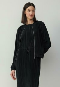 Robe noire plissée avec un col rond et une taille ajustable à cordon, accompagnée d'une veste zippée assortie avec des poignets côtelés.
