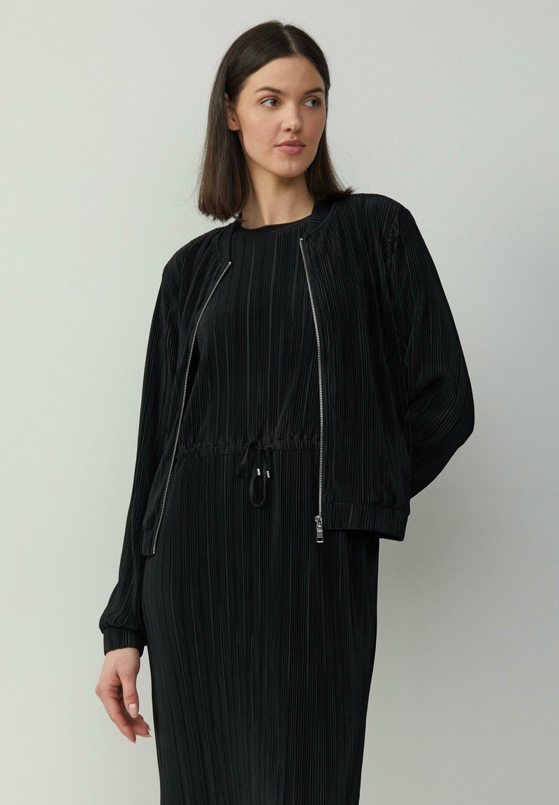 Robe noire plissée avec un col rond et une taille ajustable à cordon, accompagnée d'une veste zippée assortie avec des poignets côtelés.