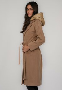 Jasnobrązowy płaszcz z paskiem w talii, z gładkim kapturem z futerka ekologicznego. Płaszcz ma dopasowany fason i boczne rozcięcia zapewniające swobodę ruchów.