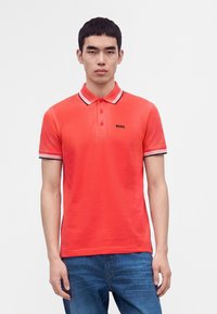Jeune homme portant un polo rouge vif avec col et manches rayés, jean bleu, debout devant un fond blanc uni.