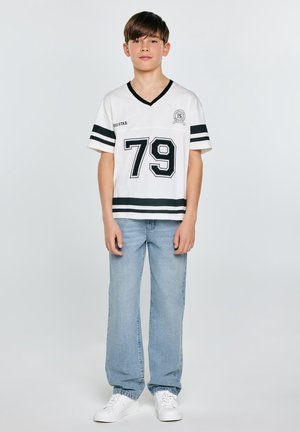 Chłopiec stojący w białej sportowej koszulce z numerem 79, jasnoniebieskich dżinsach i białych trampkach na jednolitym tle.