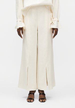 Μοντέλο που φοράει κρεμ παντελόνια με φαρδιά μπατζάκια και εμπρόσθιες σχισμές, συνδυασμένα με μπλούζα μακρυμάνικη στο ίδιο χρώμα με δεμένα μανίκια και μαύρα σανδάλια ανοιχτής μύτης.