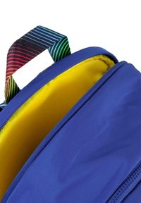 Mochila azul con un interior amarillo brillante, que presenta un asa de rayas de colores y una textura de tela suave. Apertura con cremallera.