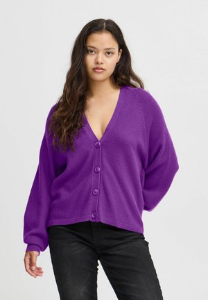 Lila Strickjacke mit tiefem V-Ausschnitt, Knopfverschluss und locker sitzenden Ärmeln, aus strukturiertem Strickmaterial. Kombiniert mit schwarzen Hosen.