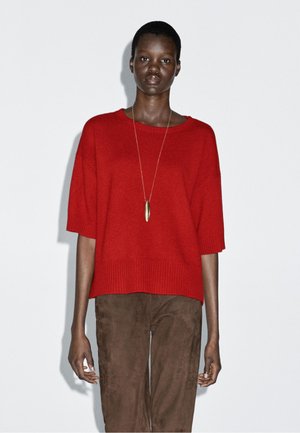 Roter gerippter Strickpullover mit lockerer Passform und kurzen Ärmeln, kombiniert mit braunen Wildlederhosen. Das Modell trägt eine lange goldene Anhängerkette.