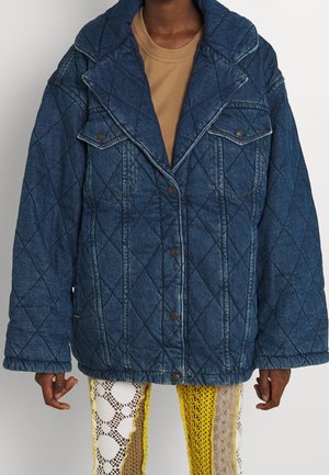 Personne portant une veste en denim bleu matelassée avec de grands revers et des boutons-pression sur un top beige, associée à un pantalon multicolore jaune et blanc en crochet.