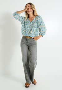 Blouse bleu clair à motifs floraux avec un décolleté en V, des manches longues et un détail à cordon, associée à un jean gris taille haute et des sandales marron.