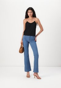 Svart plissert cami med justerbare spaghetti-stropper, kombinert med lyseblå flared jeans. Modellen holder en liten brun håndveske og har på seg beige hæler.