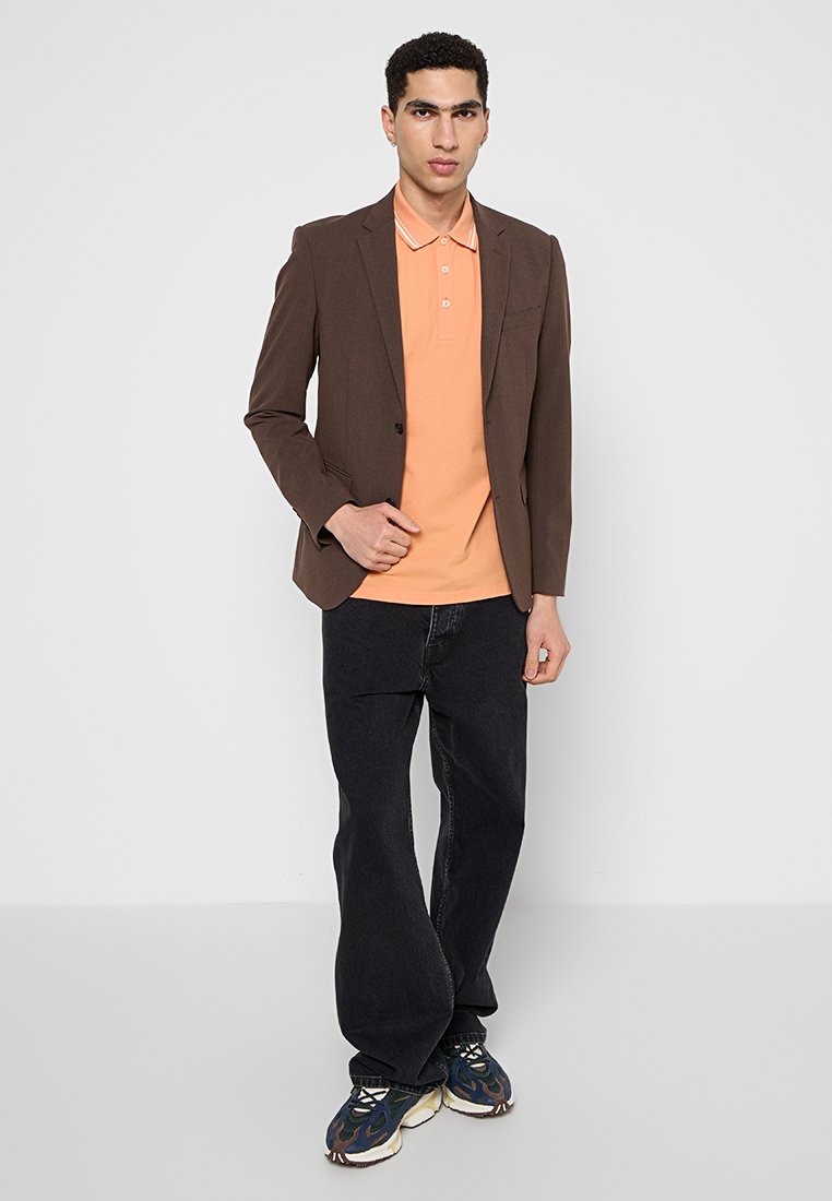 Ciemnobrązowa marynarka na pomarańczowej koszulce polo, w połączeniu z luźnymi czarnymi dżinsami i wielokolorowymi trampkami; gładki materiał o swobodnym kroju.