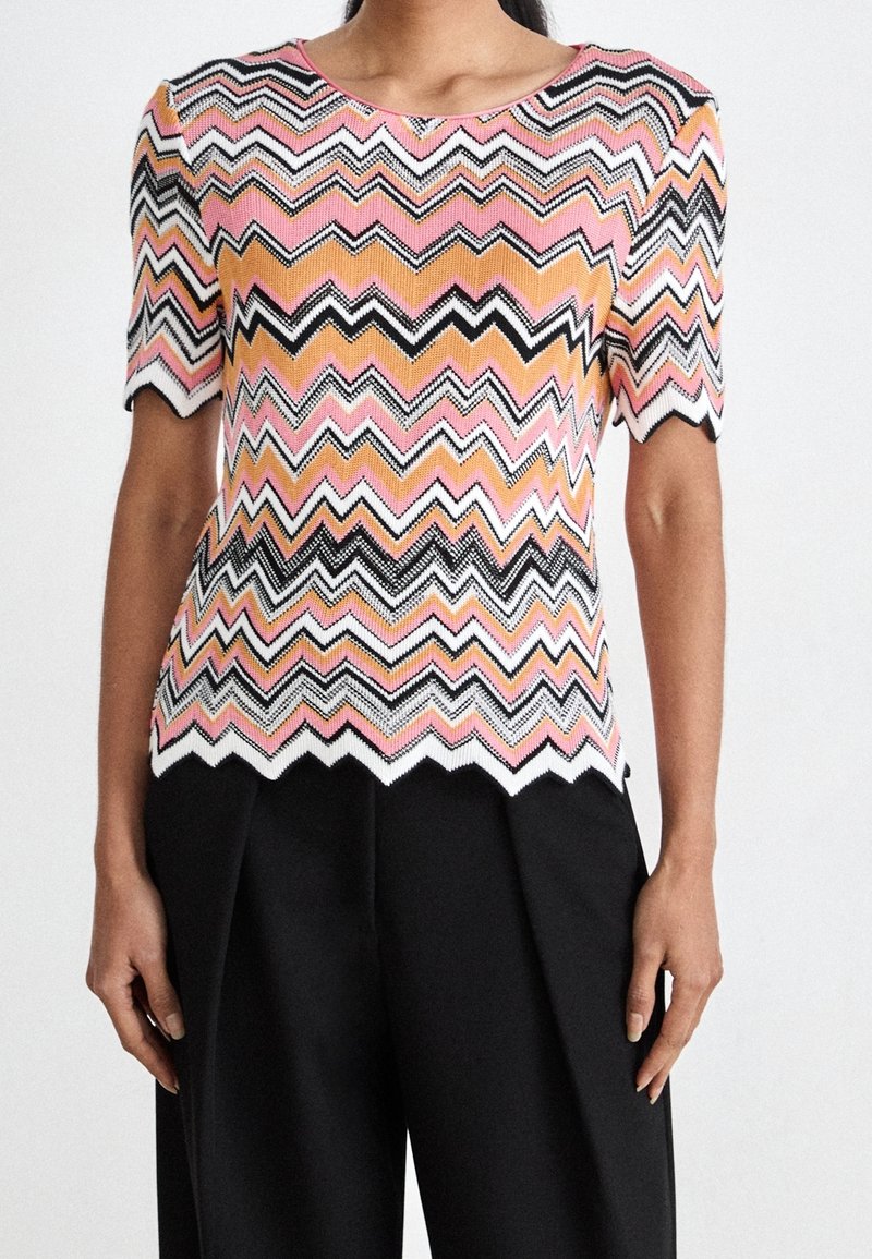 Femme portant un haut en maille à manches courtes avec des motifs en zigzag roses, orange, noirs et blancs, et un pantalon noir taille haute.