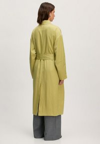 Hellgrüner Trenchcoat mit einem Gürtel, langen Ärmeln, Schlitz hinten und einer glatten Textur, kombiniert mit grauen weit geschnittenen Hosen.