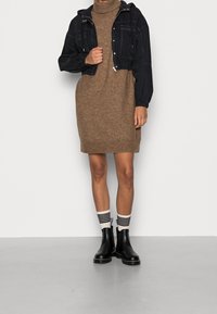 Svart kort jacka över en brun stickad klänning. Outfiten matchas med svarta ankelboots och randiga strumpor. Släta texturer och kontrasterande färger.