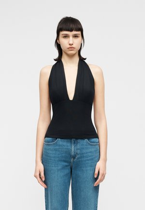 Νεαρή γυναίκα με ίσια σκούρα μαλλιά, φορόντας μαύρο τοπ με τιράντες και βαθύ V λαιμόκοψη, και μπλε ψηλόμεσο τζιν, στέκεται μπροστά σε απλό φόντο.