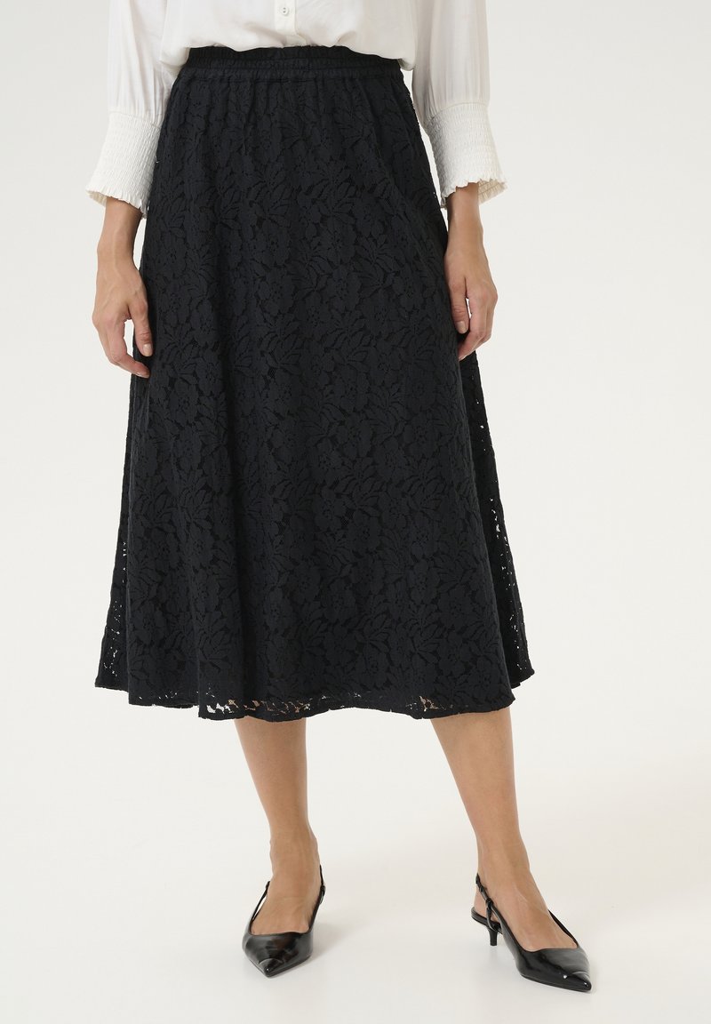 Jupe en dentelle noire longueur genou portée avec un chemisier blanc et des talons slingback noirs à bout pointu sur un fond clair.