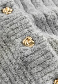 Szürke kötött szövet bordázott textúrával, fényes arany csomós gombokkal díszítve, amelyek felül hímzettek.