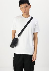 Černá obdélníková crossbody taška vyrobená z hladkého materiálu, nošená přes přiléhavé bílé tričko. Popruh je nastavitelný a vyrobený z odolné tkaniny.