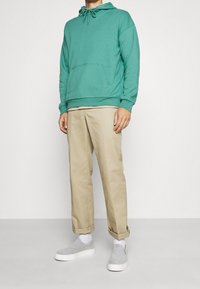 Felpa turchese con cappuccio e cordino, tasca frontale; abbinata a pantaloni beige con risvolti, scarpe grigie senza lacci e calzini bianchi.
