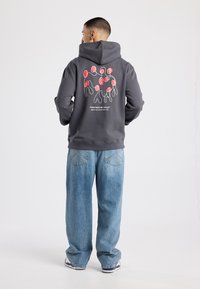 Grijze hoodie met een groot rood-wit abstract ontwerp op de achterkant, voorzien van een kangoeroezak en een verstelbare capuchon met trekkoorden. Draagt blauw spijkerbroek.