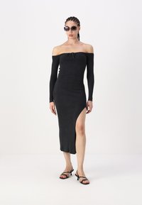 Robe noire côtelée à épaules dénudées avec des manches longues, présentant une fente latérale et un détail à nouer au niveau du col. Portée avec des sandales noires et des lunettes de soleil.