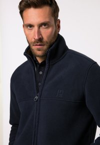 Chaqueta de forro polar navy con cuello alto, cierre de cremallera y tejido texturizado. Presenta un discreto logo bordado en el área superior izquierda del pecho.