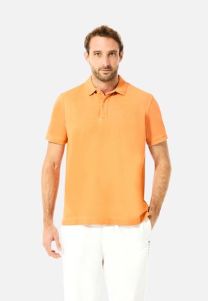 Mann trägt ein orangefarbenes kurzärmeliges Poloshirt und weiße Hosen, steht mit einer Hand in der Tasche vor einem schlichten Hintergrund.