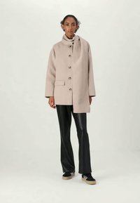 Woman wearing a beige buttoned coat with an attached scarf, black leather pants, and black sneakers with white stripes.