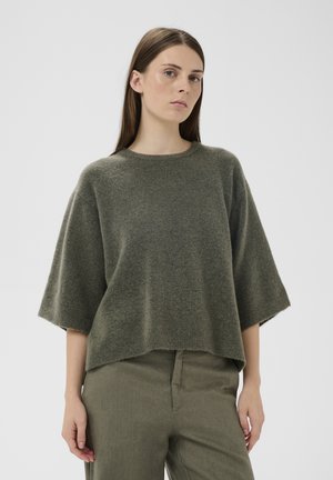 Ženska z dolgimi rjavimi lasmi, oblečena v prevelik olivno zeleni pleteni pulover in ujemajoče hlače, na enostavni svetli podlagi.