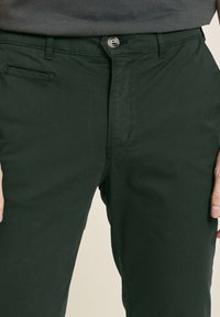 Pantaloni in cotone verde scuro con design a gamba dritta, dotati di tasca frontale, chiusura con bottone in metallo e una texture liscia.