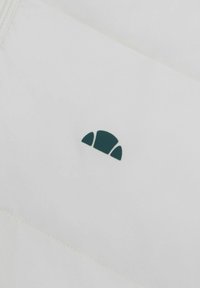 Biała tkanina z ciemnozielonym haftowanym logo przypominającym półokrąg. Tekstura jest gładka i matowa, z widocznym detalem szycia.