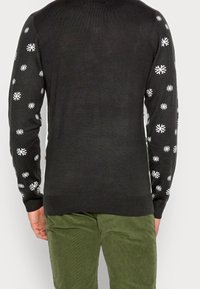 Maglione nero lavorato a maglia con motivi di fiocchi di neve bianchi lungo le maniche, polsini a coste e orlo. Abbinato a pantaloni in velluto a coste verde.