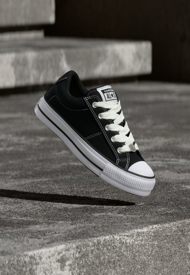 Μαύρο Converse sneaker χαμηλής κοπής με λευκά κορδόνια και σόλα, αιωρούμενο πάνω από μια επιφάνεια από σκυρόδεμα με σκάλες στο βάθος.