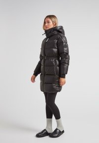Mujer de pie con un abrigo acolchado negro ceñido con cinturón, cuello alto y capucha, leggings negros, calcetines grises y zapatos negros sin cordones, sobre fondo blanco.