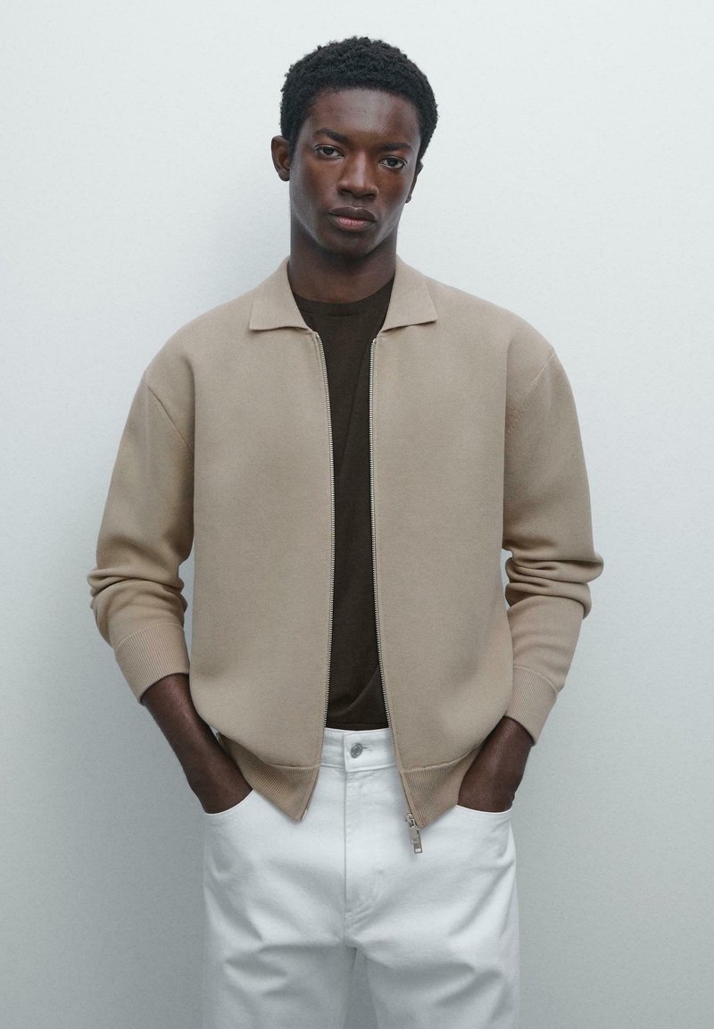 Jeune homme portant une veste beige zippée sur une chemise sombre et un pantalon blanc, les mains dans les poches, se tenant contre un fond clair.