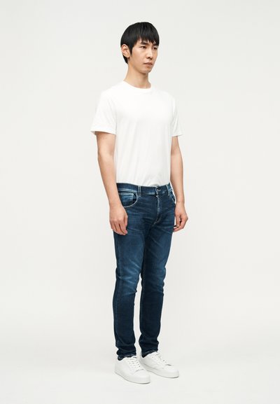 Hvid T-shirt, mørkeblå skinny jeans og hvide sneakers. Outfittet har et enkelt design og bomuldstekstur. Stående kropsholdning, neutral baggrund.