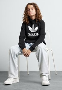 Fekete kapucnis pulóver fehér Adidas logóval, világoskék bő naci és fehér sportcipő, fehér széken ülve egy világos háttér előtt.