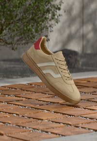 Baskets beiges avec des rayures latérales blanches et un passant de talon rouge. Présentent une surface lisse et un design profilé bas avec des fermetures à lacets.