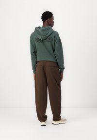 Sudadera verde con capucha y detalle del logo negro en la parte posterior, combinada con pantalones anchos marrones y zapatillas ligeras.
