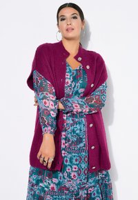 Fuchsia gestrickte Strickjacke mit großen silbernen Knöpfen, über einem mehrfarbigen, floral gemusterten Kleid mit langen Puffärmeln geschichtet.