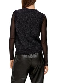 Žena v černé bezrukávové pletené vestě přes průsvitný dlouhý rukáv, s černými kalhotami z umělé kůže, zobrazená zezadu.