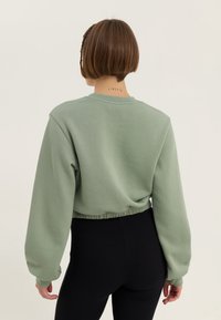 Sudadera corta verde menta con mangas largas y cinturilla fruncida, combinada con leggings negros acanalados. Vista posterior.