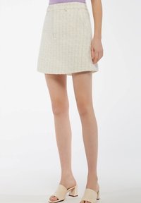 Crème mini rok van tweed met een gestructureerd patroon, voorzien van een tailleband en zakken aan de voorkant. Gepaard met open teen beige blokhak schoenen.