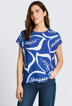 Kobieta o brązowych włosach, ubrana w niebieską bluzkę z krótkim rękawem z dużymi białymi abstrakcyjnymi wzorami liści oraz ciemnoniebieskie dżinsy, ręka w kieszeni.