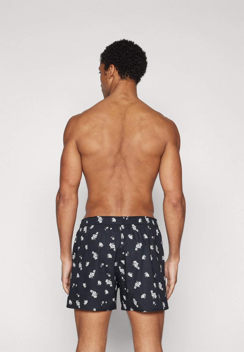 Czarne szorty kąpielowe z białym wzorem kwiatowym, elastycznym paskiem w talii i luźnym krojem, pokazane od tyłu. Gładka faktura.