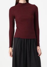 Haut long à manches en bordure bourgogne avec un col haut et un ourlet effiloché, associé à une jupe noire. Mettre l'accent sur le tissu texturé et la coupe ajustée.