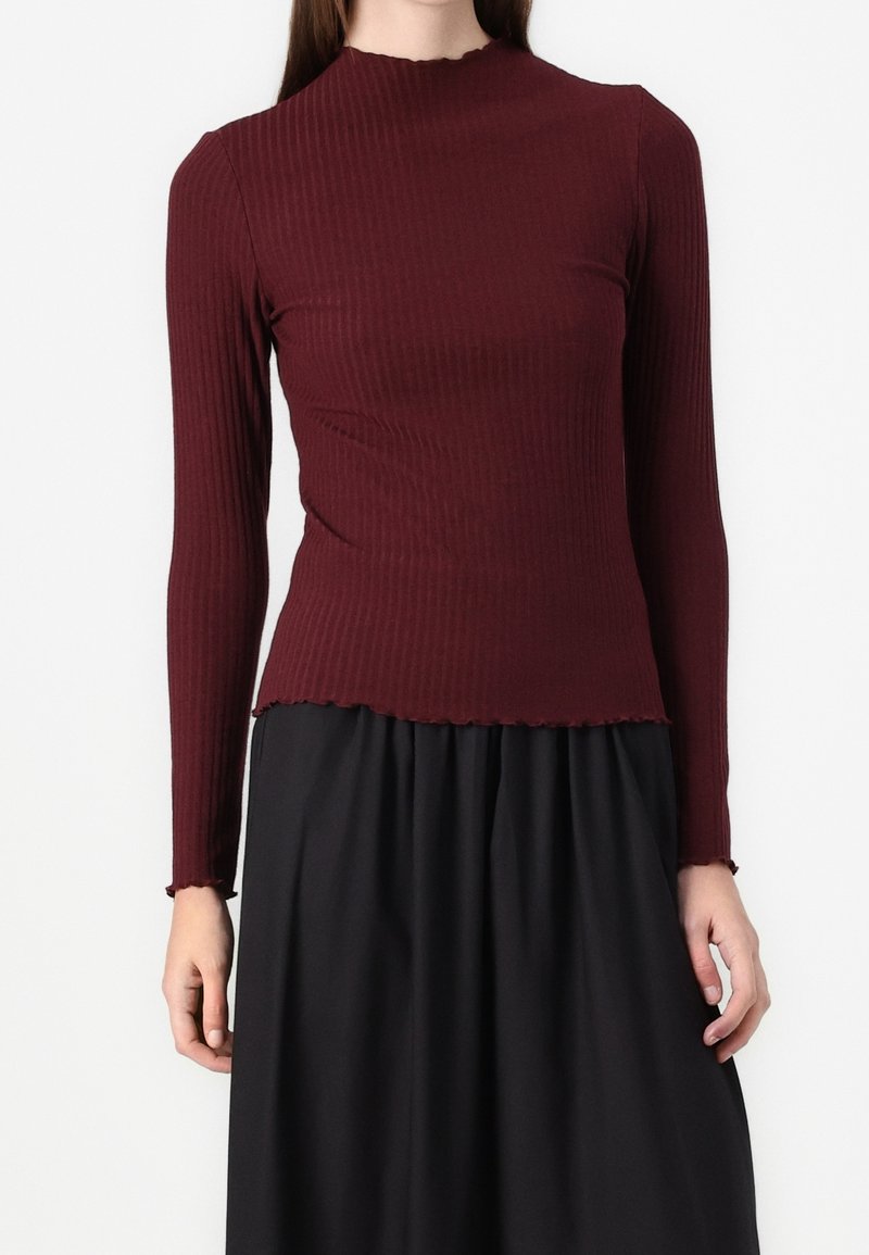 Haut long à manches en bordure bourgogne avec un col haut et un ourlet effiloché, associé à une jupe noire. Mettre l'accent sur le tissu texturé et la coupe ajustée.