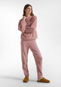 Hellrosa, flauschiges Loungewear-Set mit Rundhals, langen Ärmeln und lässig sitzenden Hosen. Die Hausschuhe sind aus braunem Wildleder mit einer weichen Innenauskleidung.