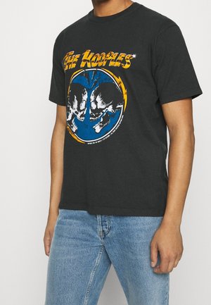 Homme portant un T-shirt noir avec un texte jaune « The Kooples » et un graphique de deux crânes opposés à l'intérieur d'un cercle bleu et jaune, associé à un jean bleu clair.