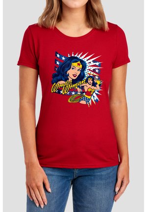 Frau trägt rotes T-Shirt mit Vintage Wonder Woman-Grafik und Text, kombiniert mit blauen Jeans vor einfarbigem Hintergrund.