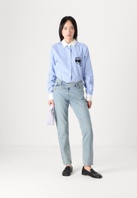 Vaaleansininen raidallinen nappipaita, jossa on valkoinen kaulus ja musta brodeeraus, yhdistettynä vaaleansinisiin farkkuihin ja mustiin loafereihin.